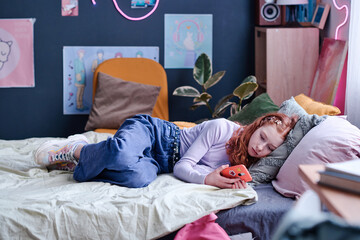 Modern Caucasian teen girl spending day indoors relaxing on bed in her bedroom watching videos in...