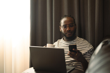 Young African American man sitting on the sofa with laptop and using a mobile phone