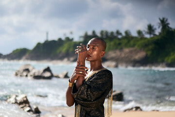 Epatage androgyne gay black man in luxury gown poses on scenic ocean beach. Non-binary ethnic...