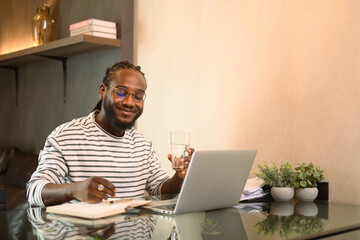 Smiling African man writing notes working remotely on laptop at home - 797553608