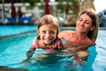 Smiling girl in pink swimsuit learns swimming in pool, mother supports. Child first swim lesson, joyful family moments. Summer day, poolside bonding, teaching kids, healthy lifestyle, water safety.