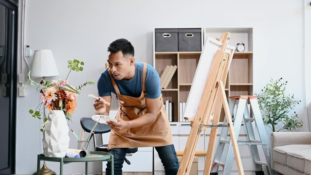 Asian male painter do artwork in art workshop, painting supplies, oil pastels, two canvas easel, creative space with paintbrush in art studio