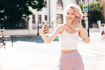 Fototapeta premium Young beautiful smiling blond hipster woman in trendy summer clothes. Carefree female posing in the street at sunny day. Positive model outdoors at sunset. Cheerful and happy. Takes selfie photos