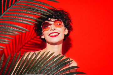 Fashion beauty portrait of young smiling brunette woman with evening stylish  makeup. Sexy model with curly short hair posing in studio. With red bright lips. Tropical palm leaf. In sunglasses