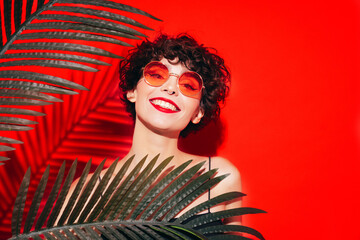 Fashion beauty portrait of young smiling brunette woman with evening stylish  makeup. Sexy model with curly short hair posing in studio. With red bright lips. Tropical palm leaf. In sunglasses