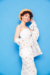 Young beautiful smiling female in trendy summer costume clothes. Carefree woman posing near blue wall in studio. Positive model having fun. Cheerful and happy. With curly short hairstyle, in hat