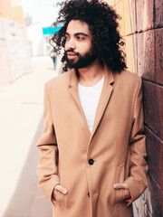 Handsome hipster model.  Unshaven Arabian man dressed in brown coat clothes. Fashion male with long curly hairstyle posing outdoors in street. Cheerful and happy. At sunny day