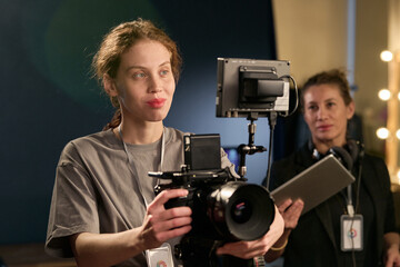 Waist up portrait of smiling young woman operating professional camera equipment on set during video production copy space