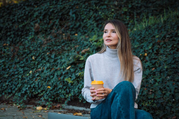A woman sits on the ground with a cup in her hand. She is wearing a gray sweater and blue jeans.