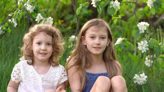 Two fair-haired girls sisters against the backdrop of a flowering apple tree