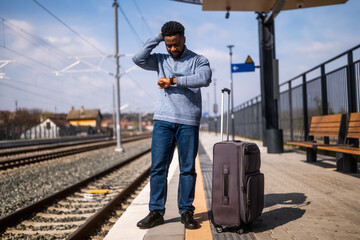 Worried man looking at his clock while standing with suitcase on a railway station.	
