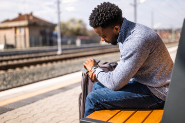Worried man looking at his clock while sitting on a bench with suitcase on a railway station.