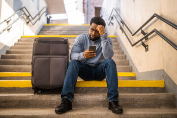 Worried man with a phone and suitcase sitting on a stairs at the railway station.	