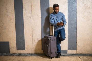Happy man with suitcase using phone while leaning against a wall in passage of the railway station. Copy space on image for your advert or text.	