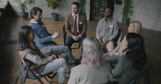 Therapist talking to multiethnic group of people sitting in circle in rehab center while men and women raising hands interacting. Therapy and rehabilitation concept.