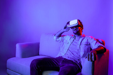 Happy man sitting at sofa while wearing VR goggle to watch funny movie. Caucasian person relaxed while looking at metaverse or virtual simulated world by using VR technology innovation. Deviation.