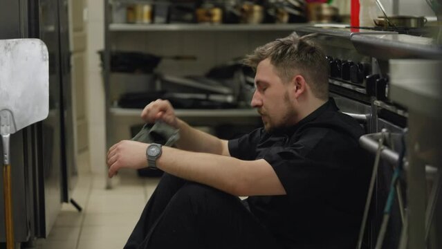Close-up of a tired male cook in a black uniform takes off his hat and straightens his hair while sitting on the floor leaning on the table in the kitchen of a restaurant