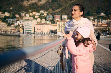 Young pretty woman and her cute daughter , a mother and little child girl in warm winter clothes, standing together on the promenade, admiring the beauty of the lake of Como in Italy