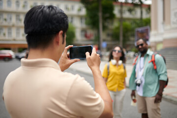 Smartphone in hands of young male tourist guide taking picture of intercultural couple standing in...