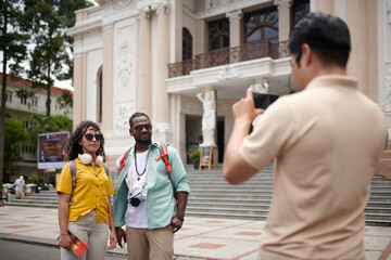 Young intercultural couple in sunglasses and casualwear standing in front of tourist guide taking picture of them against historical building