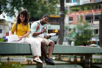 Young restful couple of travelers sitting on construction in urban environment and having takeaway...