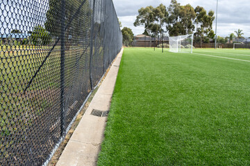 A soccer ground with artificial synthetic grass turf and water drainage gaps between mesh metal fences to allow excess water to drain away from the field and prevent pooling.