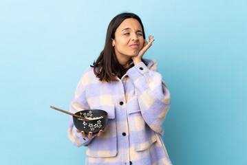 Young brunette mixed race woman holding a bowl full of noodles over isolated blue background...