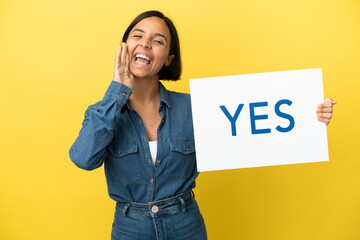 Young mixed race woman isolated on yellow background holding a placard with text YES and shouting