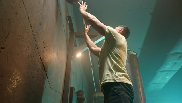 A man in a focused squat position throws a medicine ball against a wall, demonstrating strength and endurance in a gritty gym setting during an intense cross-training session. Camera 8K RAW. 