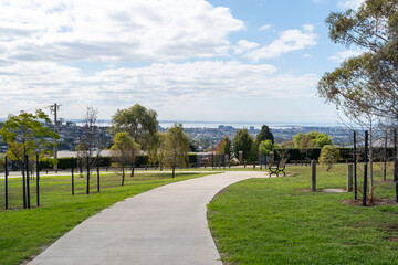 A concrete walking trail or footpath leading through a suburban neighborhood park on a hill with grass and trees with Geelong's urban skyline in the background. Wandana Heights VIC Australia.