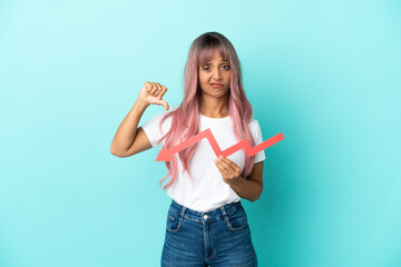 Young mixed race woman with pink hair isolated on blue background holding a downward arrow and doing bad signal