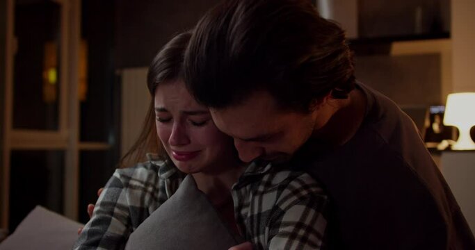A sad brunette girl in a plaid shirt sits on the sofa crying and hugs a pillow and her brunette boyfriend in a brown T-shirt supports and hugs the girl in a modern studio apartment in the evening