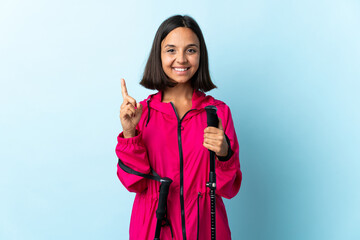 Young latin woman with backpack and trekking poles isolated on blue background showing and lifting...