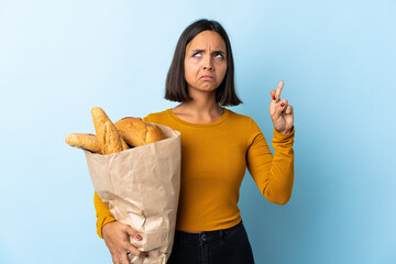 Young latin woman buying some breads isolated on blue background with fingers crossing and wishing...
