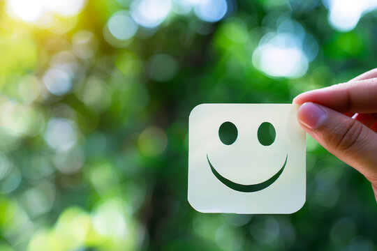 Happiness Concept with Smiley Face Card. Hand holding a card with a smiley face on it against a blurred green natural background, representing joy and positivity.