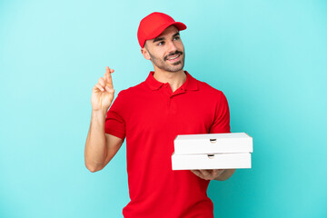 Delivery caucasian man picking up pizza boxes isolated on blue background with fingers crossing and wishing the best