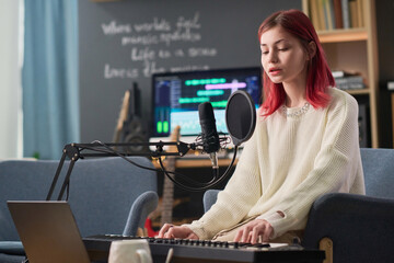 Pretty teenage girl with red hair playing electronic synthesizer by desk and singing in microphone...