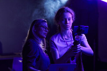 Waist up portrait of female video production crew working together and using smartphone in purple...