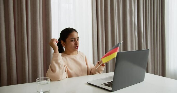 A happy brunette girl in a light jacket communicates online using a gray laptop while studying a foreign language German and shows a flag in a video conference at home online