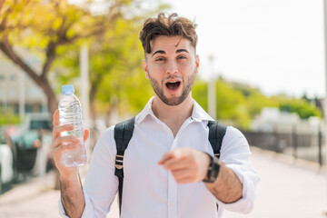 Young handsome man with a bottle of water at outdoors surprised and pointing front