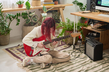 Teenage girl with red hair playing bass guitar while sitting on the floor in home studio, creating...