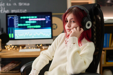Cute serene teenage girl in headphones and white sweater listening to music in armchair against...