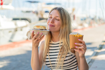 Young blonde woman at outdoors holding a burger and fried chips with happy expression