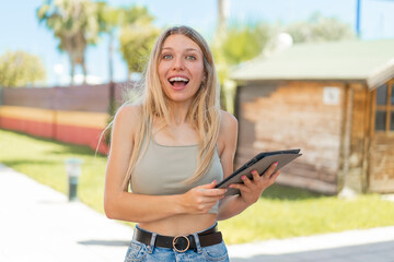 Young blonde woman holding a tablet at outdoors with surprise and shocked facial expression