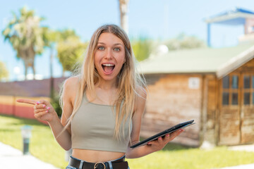 Young blonde woman holding a tablet at outdoors surprised and pointing finger to the side