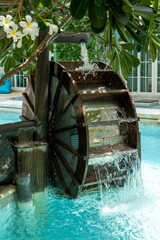 A decorative water wheel under a flowering tree through which water flows and falls into a pool of blue water in the courtyard of a condominium.