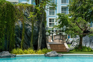 A stone pedestrian bridge with decorative lanterns and steps spans the blue water pool of a condominium at a tropical resort. Lush tropical vegetation grows around.