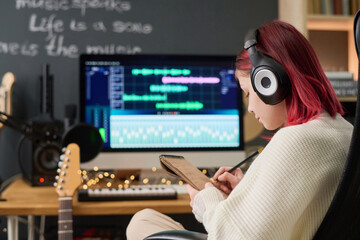 Cute teenage girl in headphones making notes in notepad while composing new music or lyrics in...