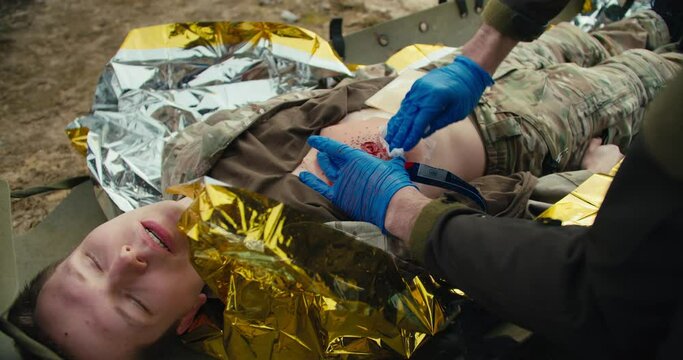 A confident male medic in blue medical gloves stops the blood of a wounded soldier in a camouflage uniform who is lying on a military medical stretcher in the combat area