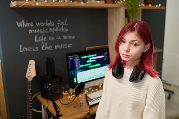 Cute serene teenager with headphones on neck looking at camera in home studio while working with...